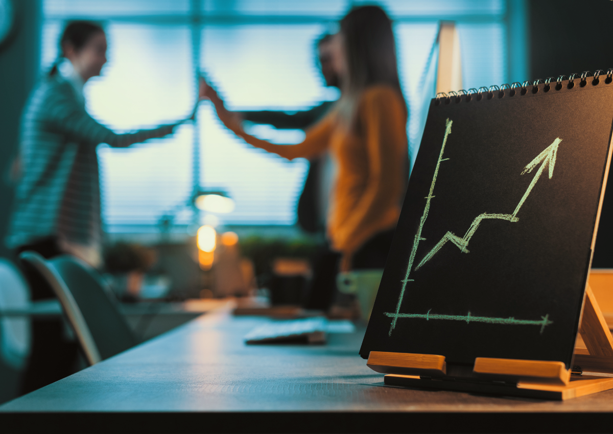 Three co workers giving a group high five in an office meeting room with a pad of paper on the table and a graph showing growth.