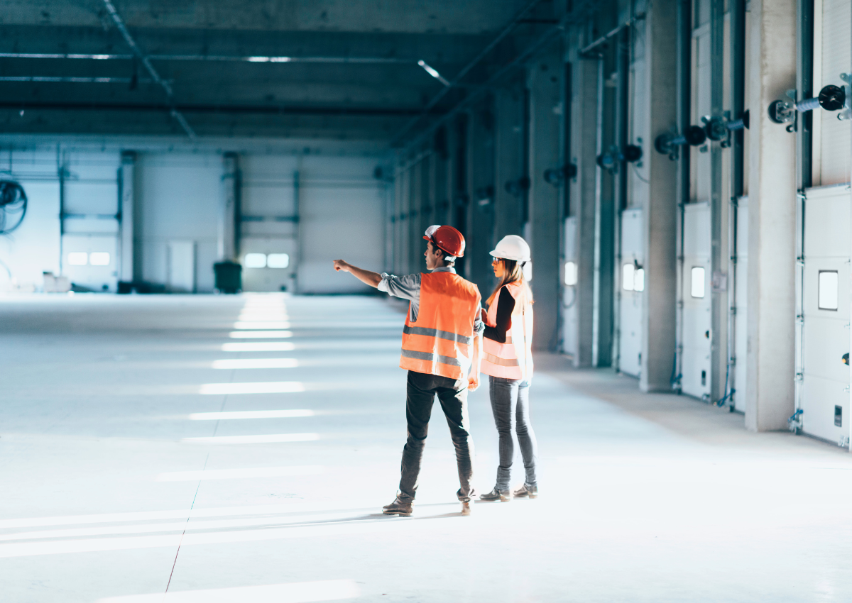 Two workers with helmets, orange reflective vests. In a warehouse. One working holding a clipboard and one pointing across the warehouse.